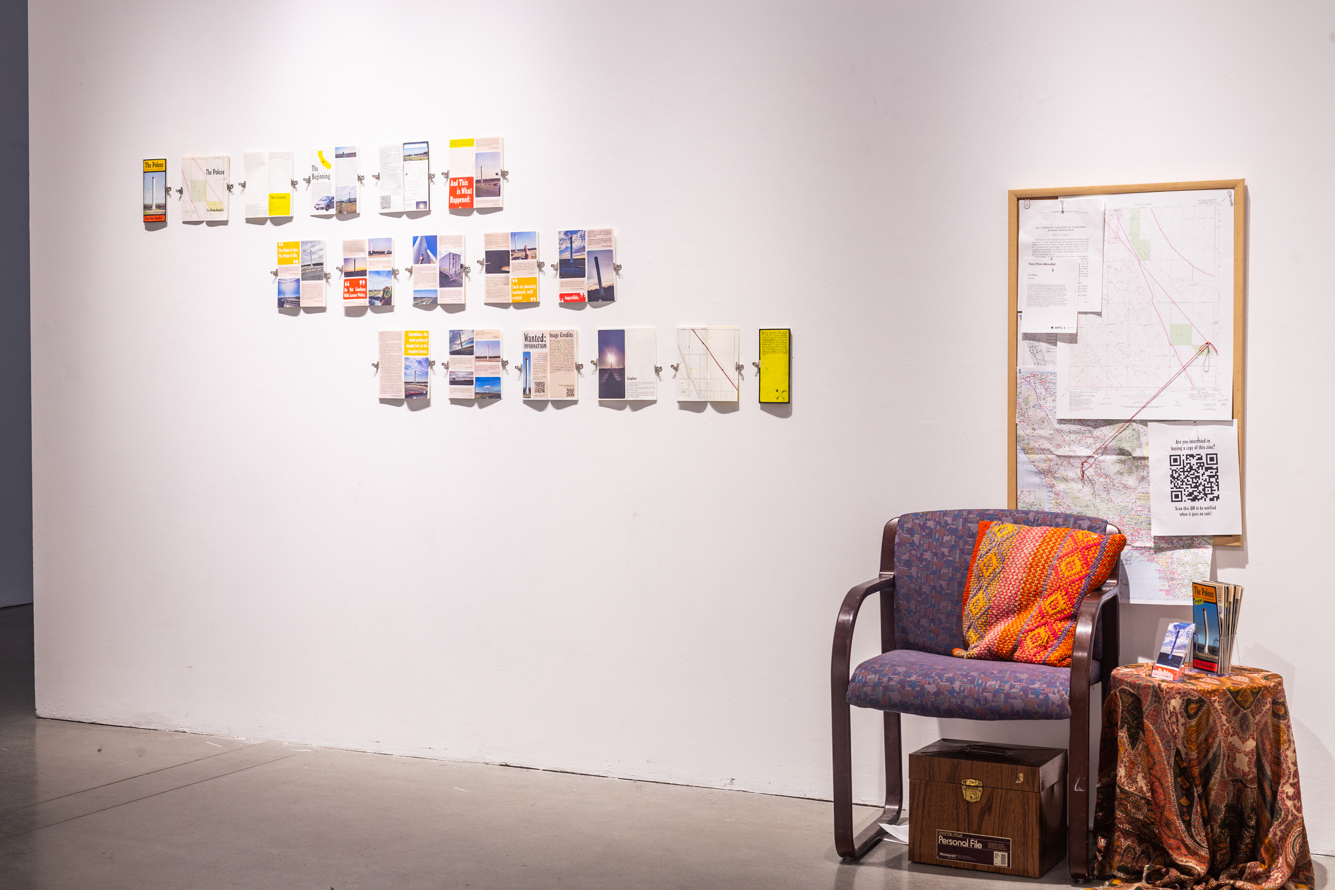 A chair sits in the center of the frame, with a pillow on it. On the right of the chair is a small circular coffee table with brochures. In the back is a white wall with a corkboard with maps and diagrams.